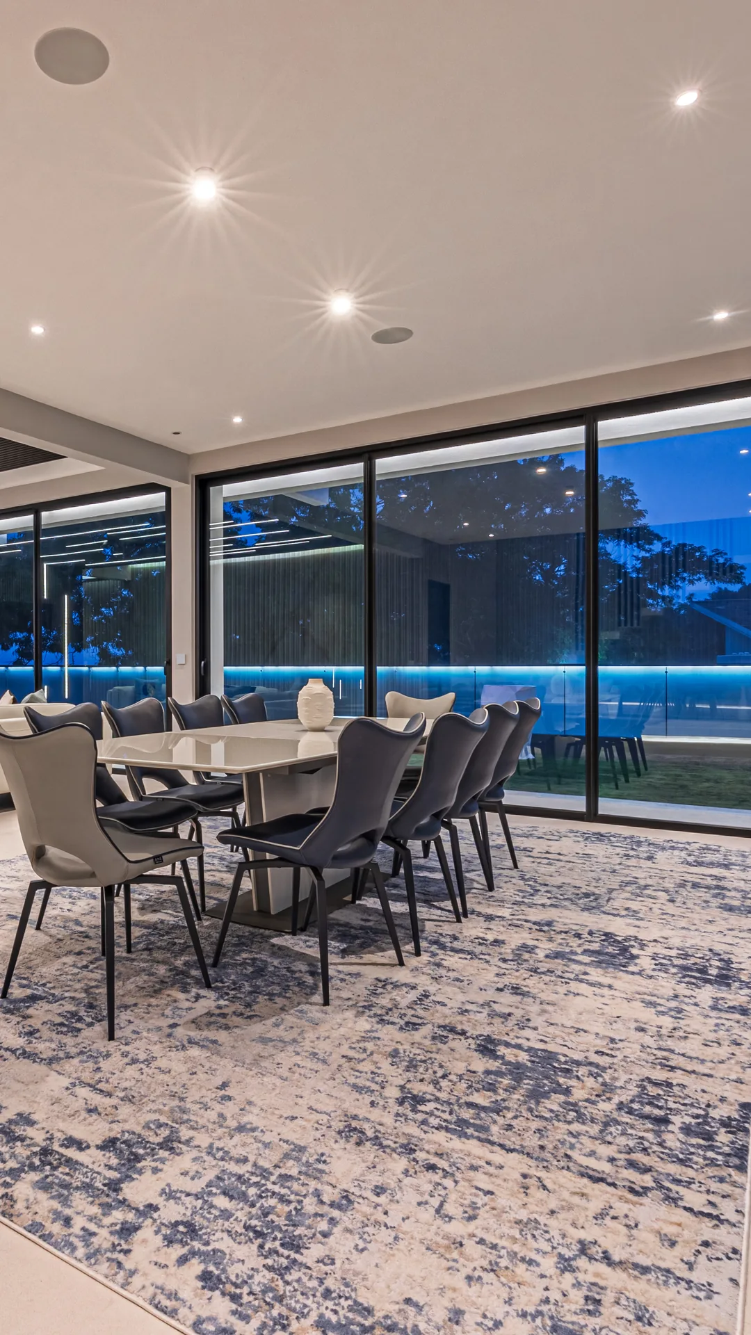 Alianz living and dining space at dusk — automated ceiling lighting, dining table and corner glazing opening to the illuminated garden