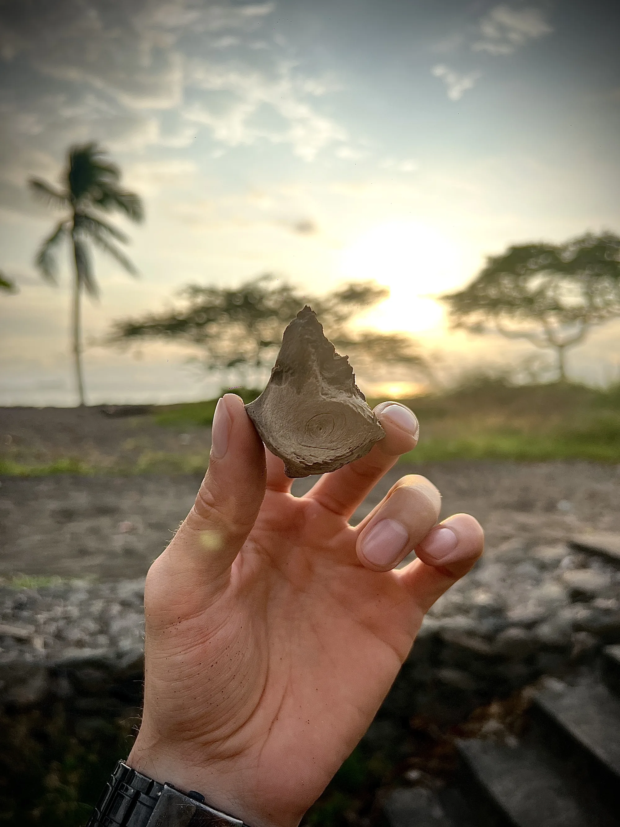 Una mano sostiene la pequeña pieza de madera con forma de ojo contra un atardecer en la playa